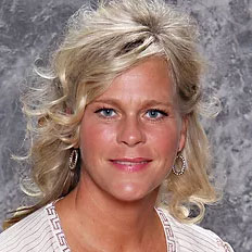 A smiling woman with shoulder-length wavy blonde hair is posing against a textured gray backdrop. She is wearing a light-colored top with a patterned trim, hoop earrings, and minimal makeup.