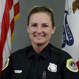 A smiling woman in a police uniform stands in front of two flags. She has light-colored hair pulled back and is wearing a dark shirt with badges and pins. The American flag is on the left, and another flag with text and an emblem is on the right.