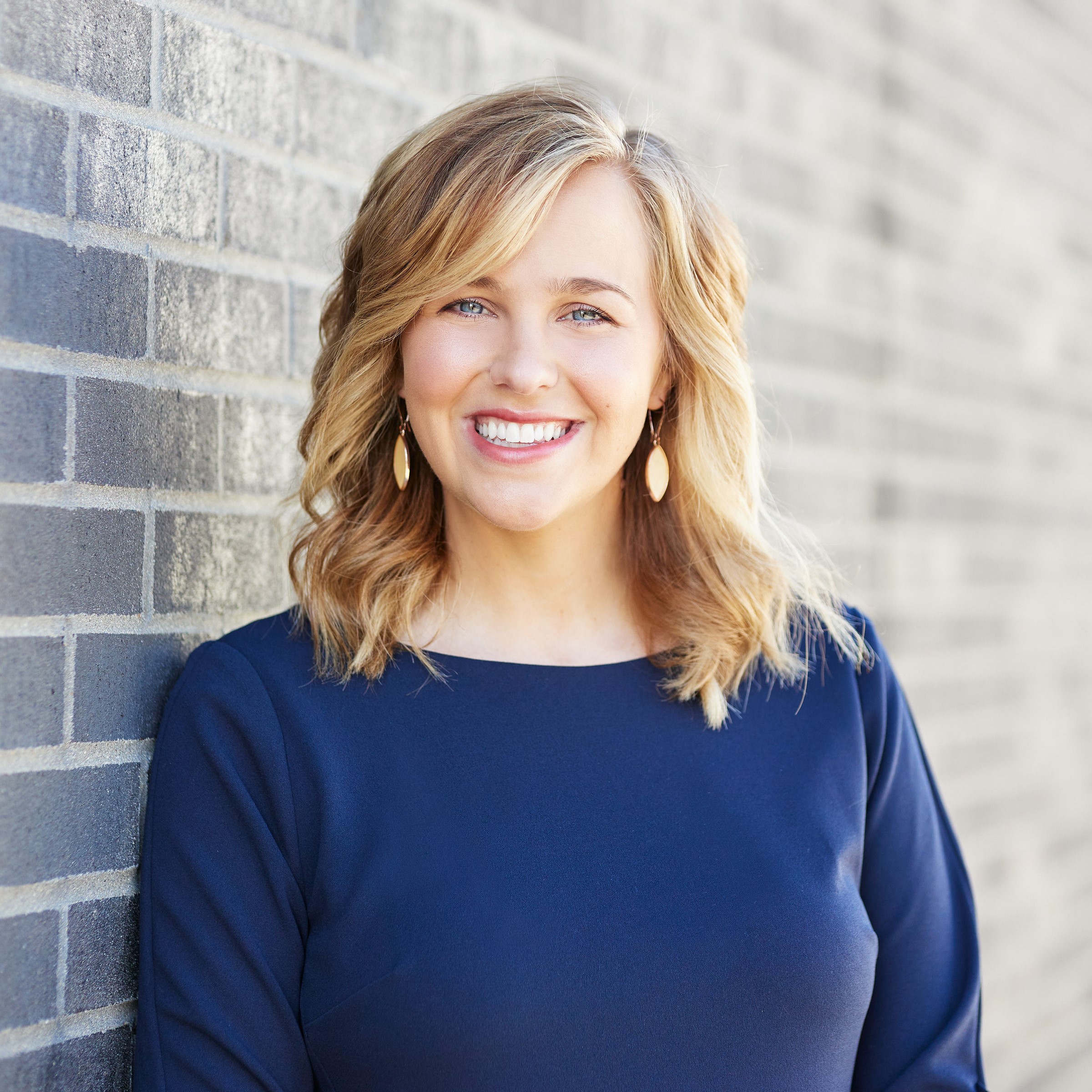 A woman with shoulder-length wavy blonde hair, wearing a dark blue long-sleeve top and gold drop earrings, smiles at the camera. She is standing against a light grey brick wall. The overall mood is friendly and inviting.