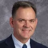 A person with short dark hair, wearing a dark suit jacket and a checkered dress shirt, smiles at the camera against a textured gray background.