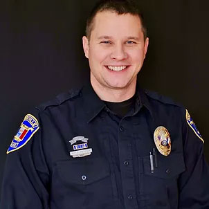 A smiling police officer in uniform stands in front of a black background. The uniform has a badge and several patches on the sleeves and chest. The officer has short brown hair.