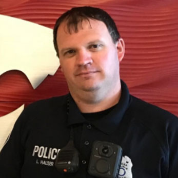 A person wearing a police uniform with a radio and body camera stands in front of a red background with a white abstract shape partially visible. The name patch on the uniform reads "L. HAUSER.