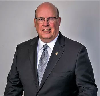 A bald man with glasses wearing a dark suit, light blue shirt, and patterned tie stands against a plain background. He is smiling and facing the camera.