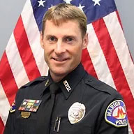 A police officer in uniform stands in front of an American flag. The officer is wearing a dark shirt with various badges and a shoulder patch that reads "Marion Police." The officer has short, light brown hair and is smiling at the camera.