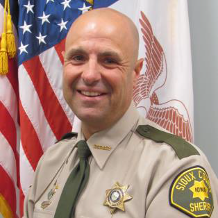 A smiling man in a sheriff's uniform stands in front of an American flag and another flag. He has a bald head and wears a beige shirt with a green tie. The badge on his uniform reads "Sioux County Sheriff" and there are other insignias on his shirt.