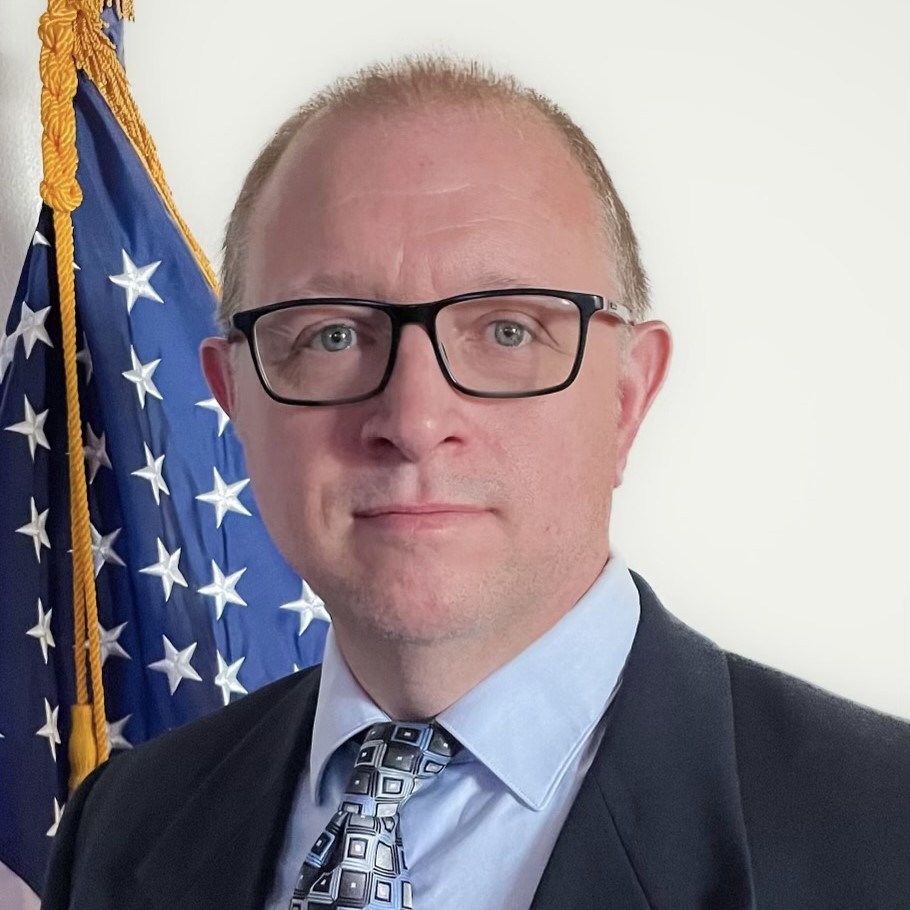 A middle-aged man with short, light brown hair and glasses is wearing a suit jacket, dress shirt, and patterned tie. He is standing in front of an American flag, which is partially visible behind him.