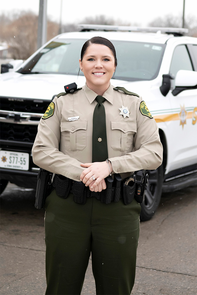 A smiling female police officer in uniform stands in front of a white police SUV on a paved surface. The officer has dark hair pulled back and wears a beige shirt, dark green pants, and a utility belt.