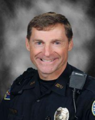 A smiling male police officer in uniform poses for a professional portrait against a gray, cloudy background. He has short brown hair, a police badge, and a radio on his shoulder.