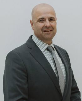 A bald man wearing a dark suit, checkered shirt, and light-colored tie smiles slightly while posing against a plain light background.